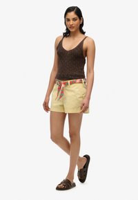 Canotta a maglia marrone, pantaloni corti gialli con cintura a fantasia floreale e sandali a slip marroni. Modella in piedi dritta su uno sfondo bianco.