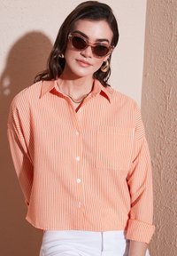 Oranje en wit gestreepte overhemd met knopen, een enkele borstzak, opgerolde mouwen en een relaxte fit. Gecombineerd met witte broek.
