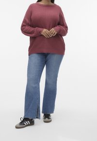 Pull en maille bordeaux surdimensionné avec un design texturé, associé à un jean évasé bleu clair avec des fentes latérales et des baskets noires avec des accents gris.