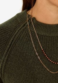 Dunkelgrüner gestrickter Pullover mit geripptem Kragen. Drei geschichtete Halsketten: eine goldene Kette mit ovalen Gliedern, eine rote Perlenkette und eine goldene Kette.