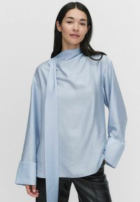 Vaaleansininen pitkähihaisen paita, jossa on korkea kaulus ja sivusiteen yksityiskohta. Valmistettu sileästä kankaasta, leveät hihansuut ja löysä istuvuus.