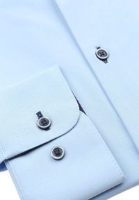 Lichtblauw button-up shirt met een gestructureerde stof, voorzien van donkere knopen en contrasterende details aan de binnenkant van de mouwen. De mouwen zijn op maat gemaakt en afgerond.
