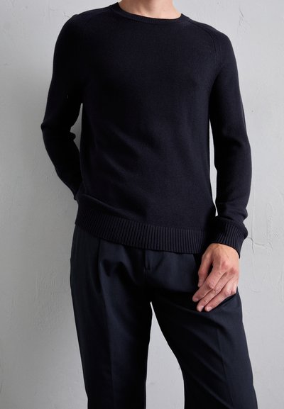 Homme portant un pull noir à col ras du cou côtelé et un pantalon noir se tenant contre un mur texturé clair.