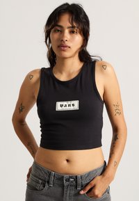 Crop top in cotone nero con scollatura rotonda e etichetta ricamata "Vans" su una toppa rettangolare. La modella indossa jeans grigi e ha tatuaggi visibili.