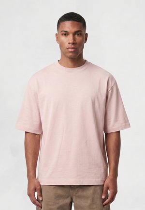 Jeune homme aux cheveux courts portant un t-shirt oversize rose clair à manches courtes et un pantalon beige, debout devant un fond clair uni.