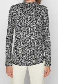 Marineblauwe bloemenblouse met lange mouwen en hoge hals, voorzien van witte bloemenpatronen en een zachte stofstructuur. De voorkant is iets langer.