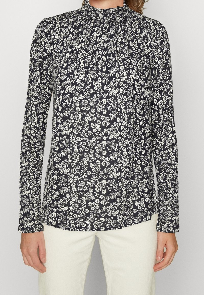 Marineblauwe bloemenblouse met lange mouwen en hoge hals, voorzien van witte bloemenpatronen en een zachte stofstructuur. De voorkant is iets langer.