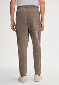 Man in taupe gemusterde joggerbroek met zijstrepen en witte sneakers, staand tegen een effen lichte achtergrond.