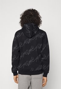 Schwarzer Hoodie aus strukturiertem Stoff mit einem wiederholten grauen Schriftmuster. Er verfügt über eine Vordertasche und eine Kapuze mit Kordelzug.