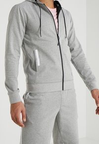 Hoodie gris con cremallera y cordones negros, que presenta un bolsillo lateral con un acento blanco, hecho de tejido suave. Lleva puesto una camiseta rosa clara.