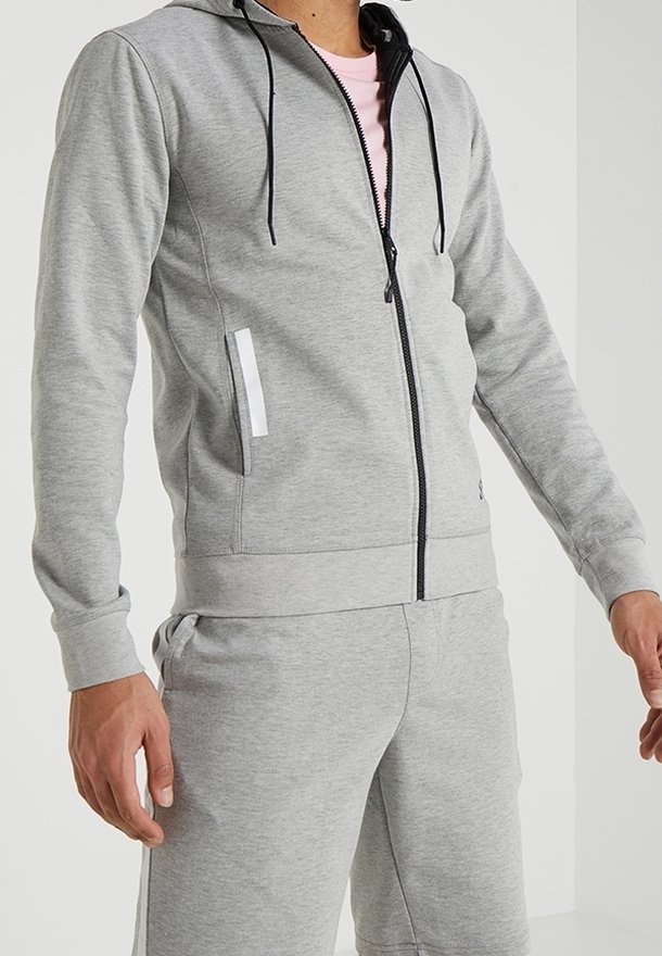 Hoodie gris con cremallera y cordones negros, que presenta un bolsillo lateral con un acento blanco, hecho de tejido suave. Lleva puesto una camiseta rosa clara.