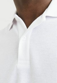Polo shirt blanc en tissu texturé, présentant un petit motif à carreaux, un col et deux boutons près du décolleté.