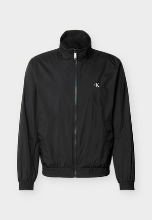 Must värvi kerge lukuga jakk, millel on kõrge krae, elastsed mansetid ja äär, ning vasakul rinnal väike valge "cK" logo.