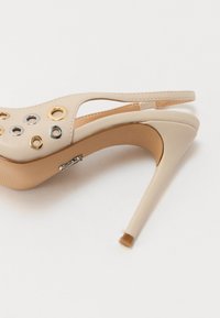Beige high-heeled shoe with an open design, featuring gold and silver grommets, a sleek silhouette, and a narrow stiletto heel.