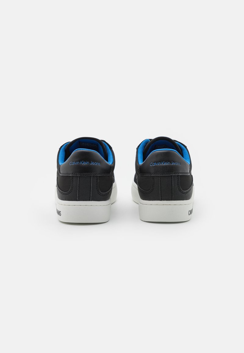 Baskets noires avec des accents bleus, dotées d'un matériau lisse, d'un design bas, d'un col rembourré et d'une semelle en caoutchouc blanche. Logo visible sur le talon.