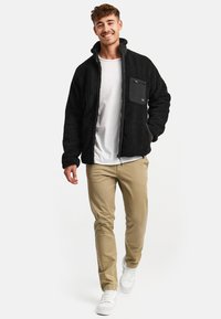 Black fleece jacket with a zippered chest pocket, textured material, high collar, and front zipper, paired with khaki pants and white sneakers.