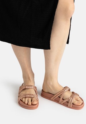 Feet wearing rose gold braided strap sandals and a black textured dress with a side slit on a white background.