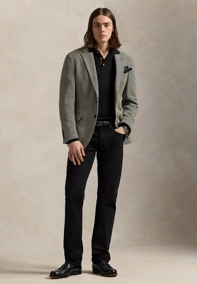 Jeune homme aux cheveux bruns mi-longs portant un blazer gris à chevrons, un polo noir, un pantalon noir et des chaussures noires, debout avec une main dans la poche.