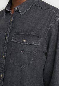 Camicia in denim grigio scuro con maniche lunghe, chiusura a bottoni sul davanti, due tasche sul petto e bottoni metallici. Presenta un piccolo logo sul taschino.
