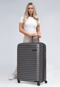Valise rigide grise avec des rainures verticales, poignée rétractable et quatre roues pour la mobilité. Dispose d'une fermeture éclair et d'un logo.