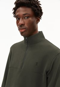 Pullover mit Viertelreißverschluss in dunkelolivgrün, aus weichem Stoff. Verfügt über ein tonales Logo und eine glatte Textur mit einem hohen Kragen.