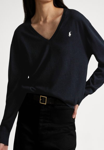 Navy V-neck sweater made from soft material, featuring a small white logo on the chest, paired with high-waisted black trousers and a gold belt.