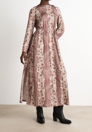 Person wearing a long-sleeve, floor-length floral and paisley patterned dress with a waist tie, paired with black heeled ankle boots.