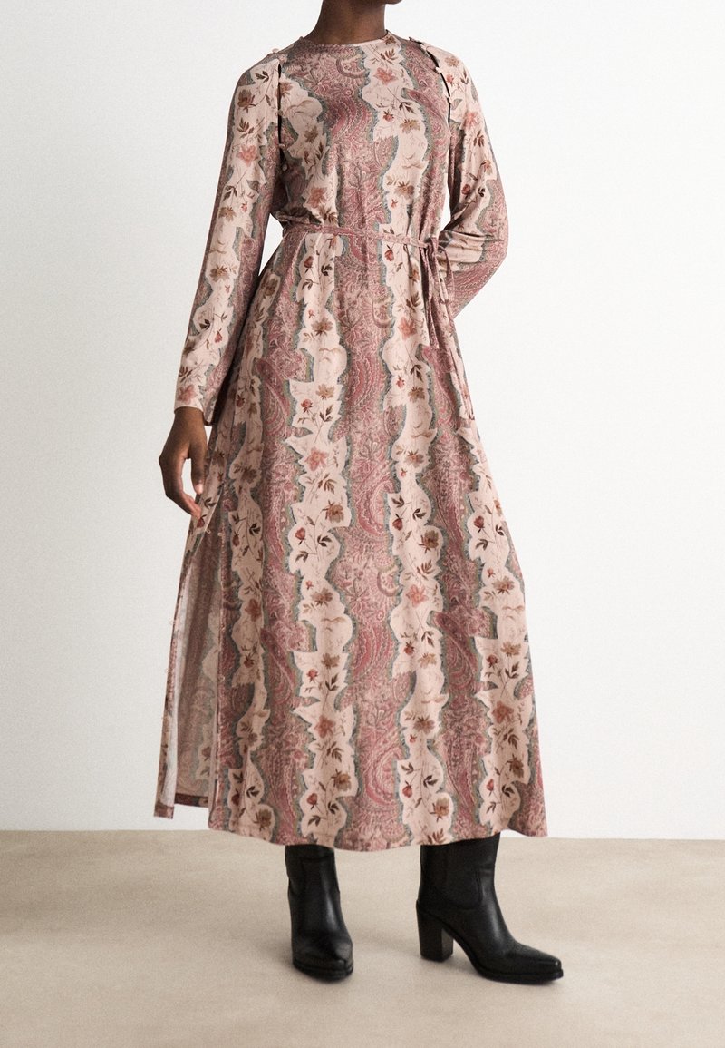 Personne portant une robe longue à manches longues, aux motifs floraux et paisley, avec une ceinture à la taille, associée à des bottines noires à talons.
