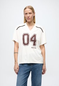 T-shirt en jersey léger crème à manches courtes, col en V, imprimé bordeaux "04" et logo "MODSTRÖM" en dessous. Porté avec un jean bleu.