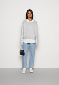 Un sweat gris avec un col blanc, superposé sur un haut clair. Porté avec un jean bleu clair et des baskets blanches, tenant un petit sac noir.