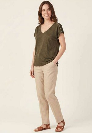 Femme debout, portant un haut à manches courtes col en V vert olive, un pantalon beige et des sandales marron, souriante avec une main dans la poche.