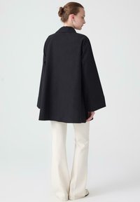 Black, loose-fit coat with wide sleeves and a back vent, paired with white flared pants. Minimalist design with no visible embellishments.