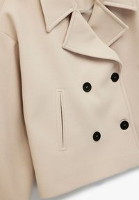 Beige dobbelknappeljakke med store sorte knapper, slagkrave, et tekstureret stof og sidelomme-detaljer.