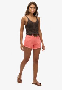 Pantalones cortos de mezclilla coral brillante con dobladillo deshilachado, combinados con una blusa de crochet oscura. Llevados con un cinturón marrón y sandalias marrones tipo slide.