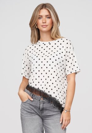 Ung kvinde med blondt hår iført en hvid kortærmet prikket top med sort blonderkant og grå jeans, poserer med den ene hånd i lommen.