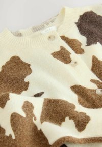 Krämfärgad koftan har ett brunt mönster med kor, ribbad halsringning och knäppning med knappar. Mjukt, texturerat ulltyg med släta detaljer.