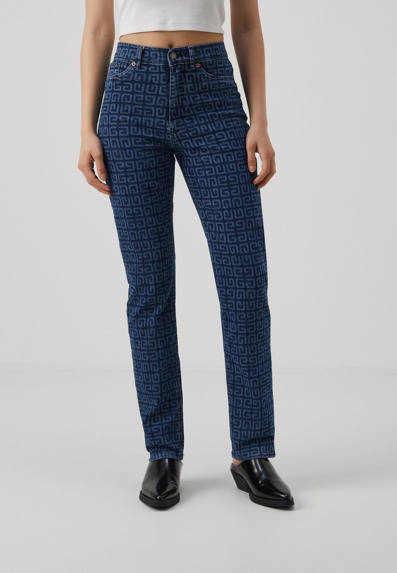 Femme portant un jean slim taille haute bleu avec un motif géométrique et des chaussures noires à talons, debout avec les mains détendues le long du corps.