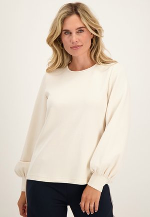 Blonde vrouw met golvend haar draagt een crèmekleurige longsleeve met opgeblazen manchetten en een zwarte broek, staand voor een effen lichte achtergrond.