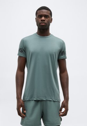 Noor mees, kes kannab sobiva värvitooniga tuhmi rohelist sportlikku T-särki ja pükse, seisab paigal ühetaolise hele tausta ees.