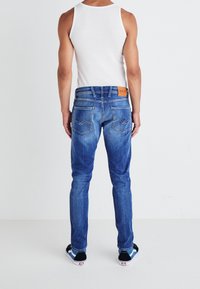 Modré džíny v slim fit s koženou nášivkou na pase a výrazným prošíváním zadních kapes. Nosí se s bílým topem bez rukávů.