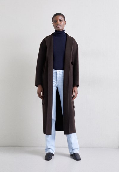 Manteau long marron avec une coupe décontractée, poches latérales et devant ouvert, porté sur un col roulé navy et un jeans bleu clair. Des chaussures noires complètent le look.