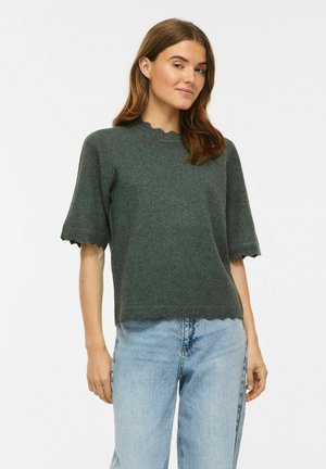 Femme aux cheveux bruns portant un pull vert foncé à manches courtes avec des bords festonnés et un jean bleu clair sur un fond uni.