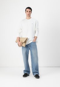 Hvit hettegenser, lyseblå vide jeans, svarte sko, holder en brettet beige veske med stropp. Minimalistisk design, enkle teksturer.