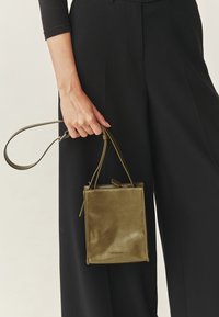 Olijfgroene leren handtas met een gladde textuur, rechthoekige vorm en korte handgreep, vastgehouden door een hand. Eenvoudige stiksels en logodetail zichtbaar.