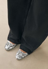 Ezüst flitteres cipők hegyes orral, masnis díszítéssel, széles fekete farmerral viselve, semleges háttér előtt bemutatva.