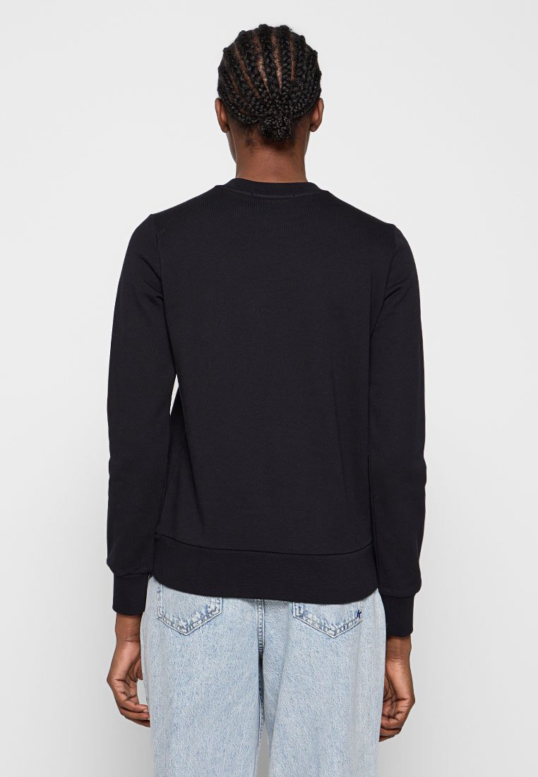 Eine Person mit geflochtenem Haar, die ein schwarzes langärmliges Shirt und eine hellblaue Jeans trägt, steht mit dem Rücken zu einer schlichten Hintergrundwand.