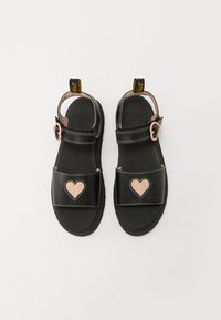 Svarte sandaler laget av syntetisk materiale med flat såle. Har to stropper, sirkulær spenne og et rosa glitrende hjertepryd på hver fotseng.