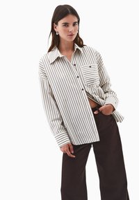 Junge Frau mit langen braunen Haaren, die ein übergroßes weißes Hemd mit schwarzen vertikalen Streifen und dunklen weiten Hosen trägt, mit der Hand in der Tasche.