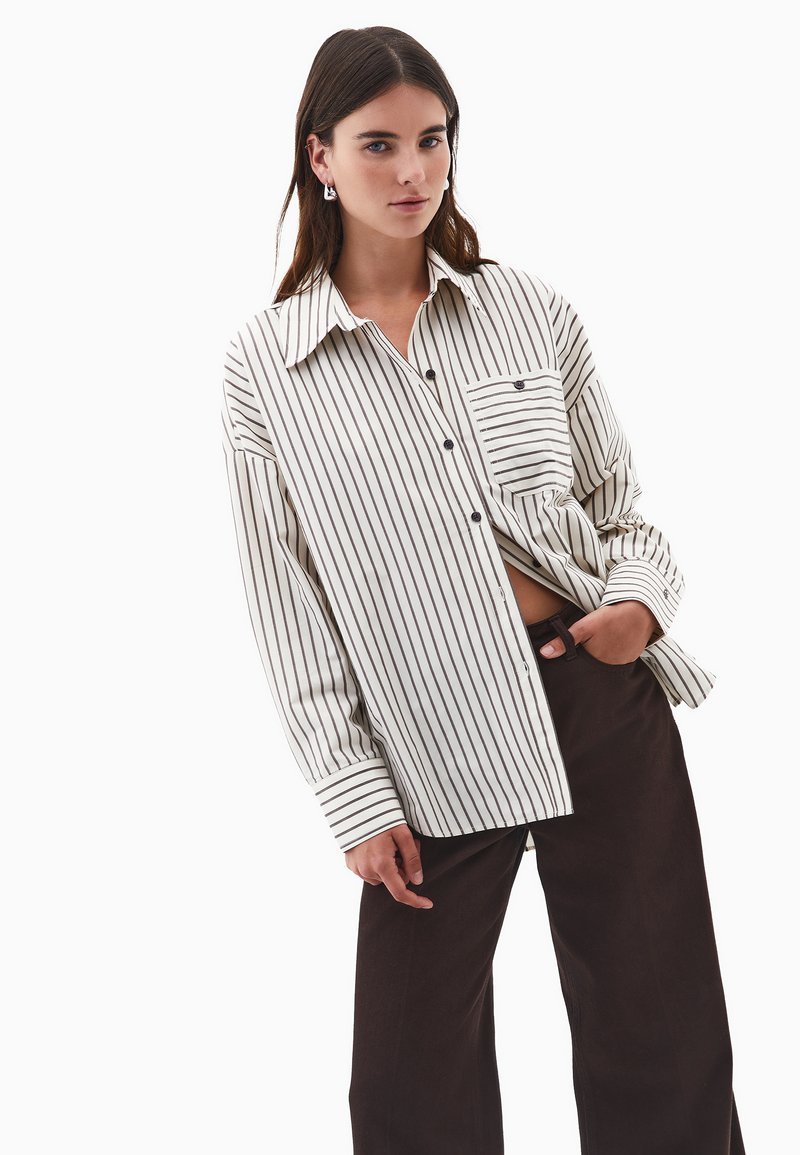 Junge Frau mit langen braunen Haaren, die ein übergroßes weißes Hemd mit schwarzen vertikalen Streifen und dunklen weiten Hosen trägt, mit der Hand in der Tasche.