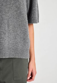 Rövid ujjú, szürke kötött pulóver bordás szegéllyel. A textúrált anyag kontrasztban áll a sima, olívazöld nadrággal, amely zsebadatot mutat.